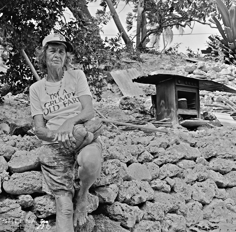 Erlene Downie and her stove to heat water.jpg - Erlene has no water or electricity. She gets water from a standpipe down the road and uses a kerosene lamp for light. She heats water once a week on this outside stove to have a wash.